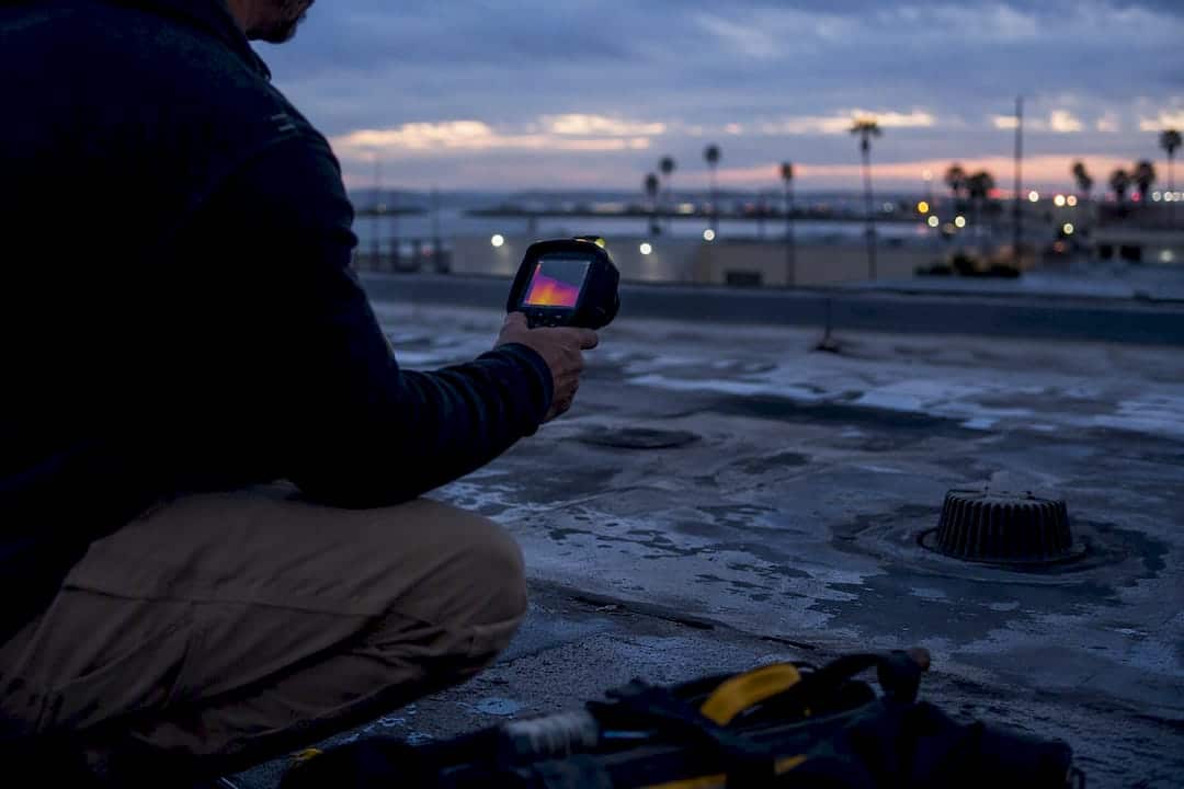 Inspector scanning flat roof with infrared camera at dusk
